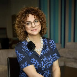 Zdjęcie Izabeli Walczewskiej-Schneyder, kobiety z curly hair i okularami, uśmiechniętej, w niebieskiej bluzce z wzorem, na tle nowoczesnego wnętrza.