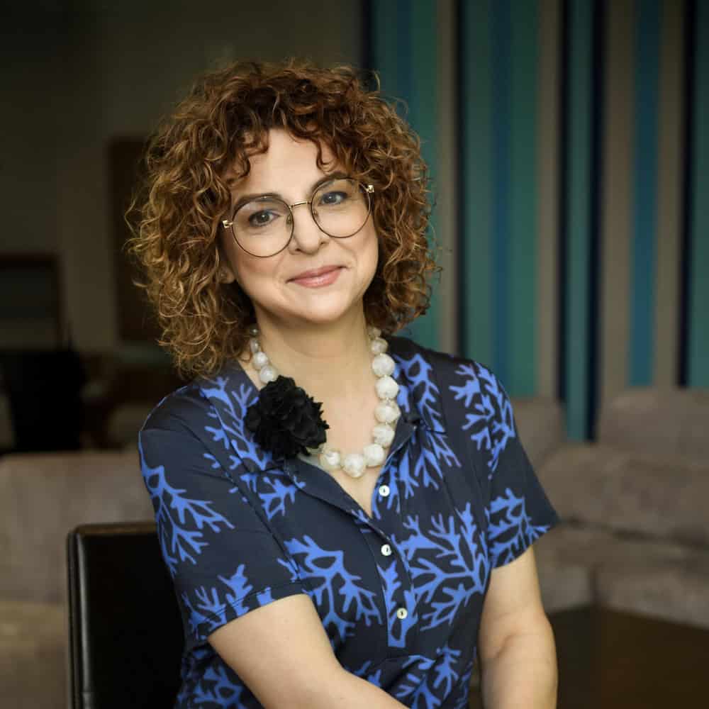 Zdjęcie Izabeli Walczewskiej-Schneyder, kobiety z curly hair i okularami, uśmiechniętej, w niebieskiej bluzce z wzorem, na tle nowoczesnego wnętrza.